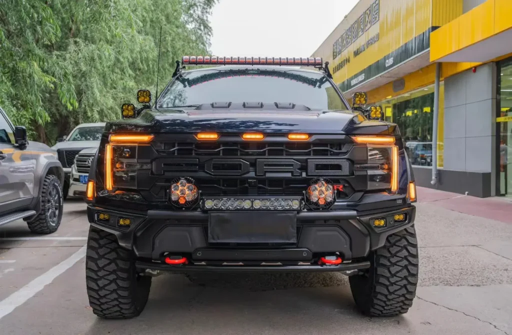 Ford Raptor outside a custom shop with grill lights installed, shown alongside auxiliary spotlights, fog lights, and projector headlights.
