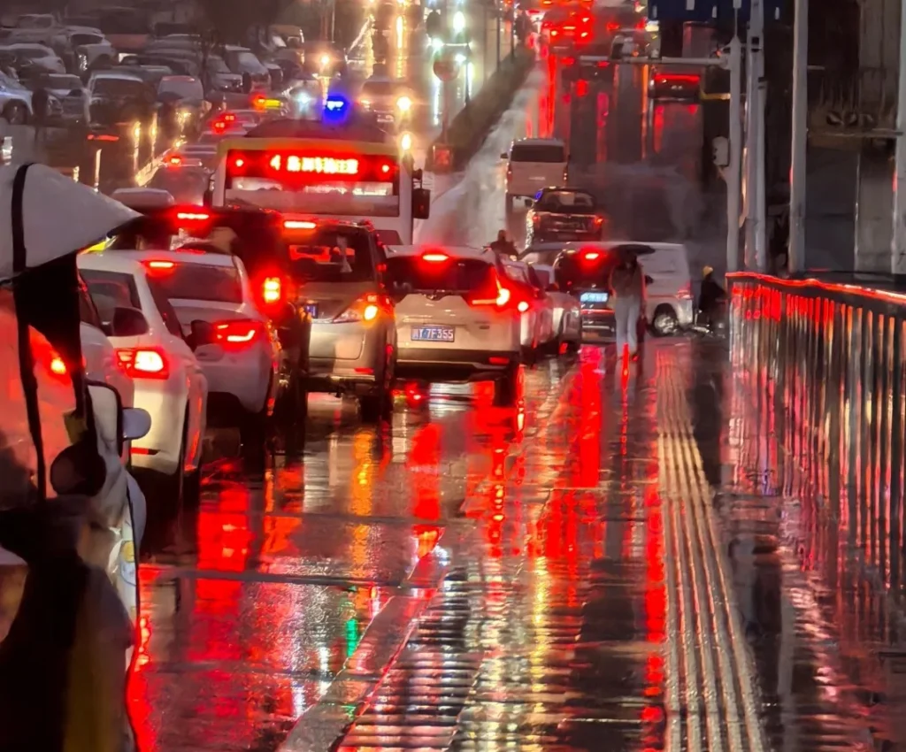 Rear-view driving scene on a rainy night; red tail/brake light glow reflects on wet asphalt and stretches into a long smear, showing how glare and bloom can make rear fog lights and brake lights harder to distinguish.