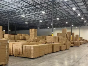 Cartons de phares de projecteurs OEM stockés sur des palettes dans une zone de rayonnage d'entrepôt avec des zones de manutention claires, des allées pour chariots élévateurs et des marquages d'orientation des cartons.